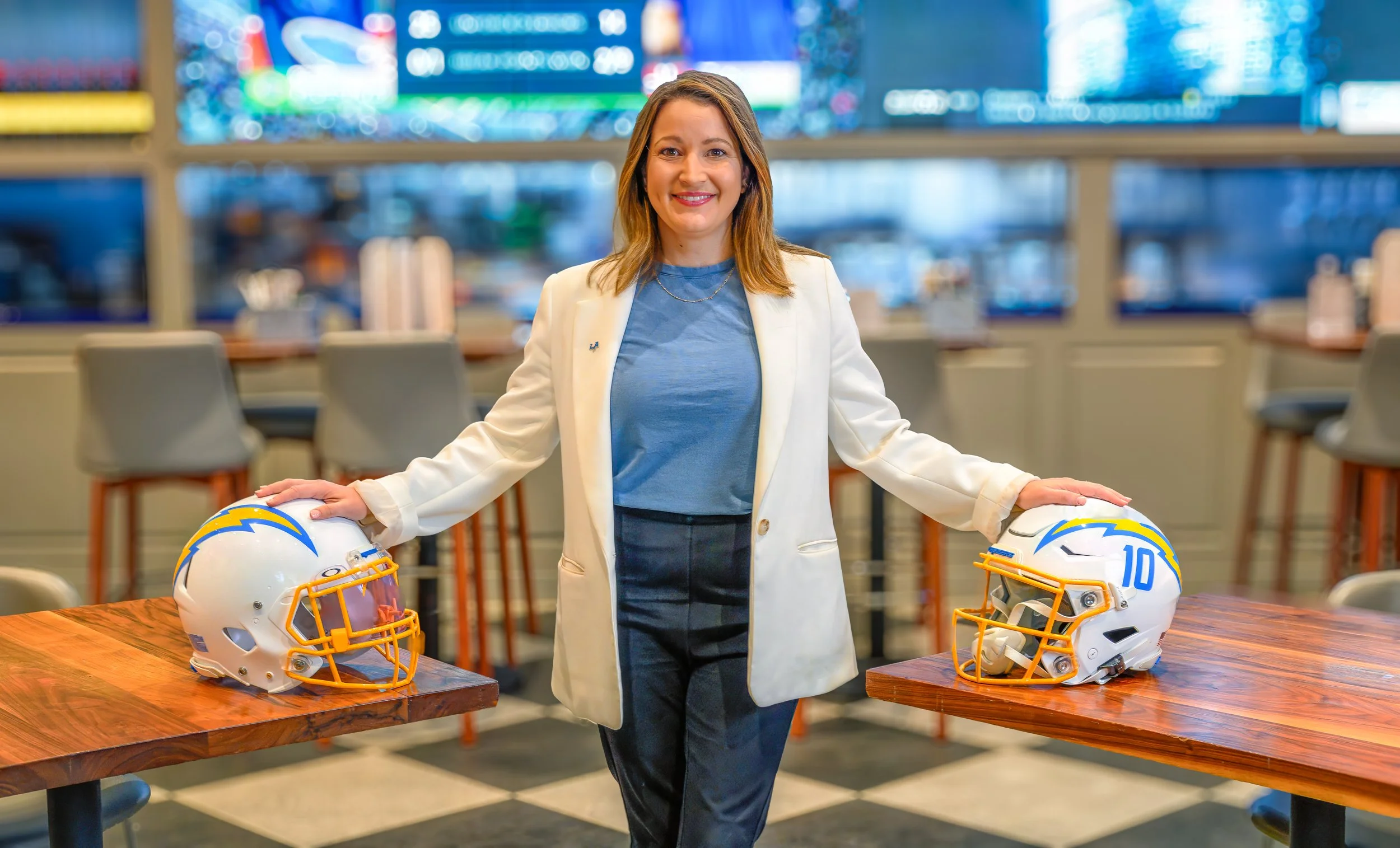 A woman in business attire smiling at the camera, standing between two tables with football helmets from the Los Angeles Chargers on either side, in a brightly lit room with colorful screens in the background.