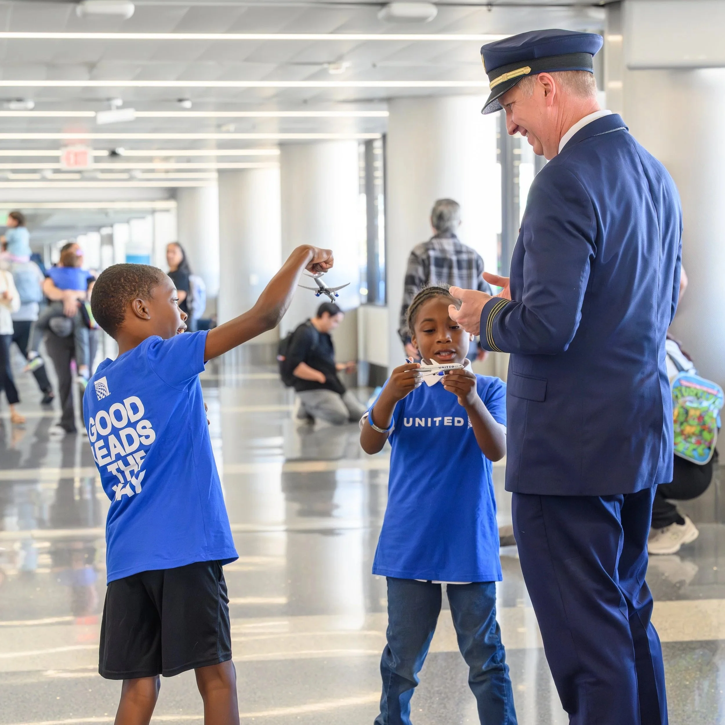 A female airline pilot in uniform smiling and talking with children at an airport gate. One child is holding a toy airplane. The children are wearing blue shirts with the words 'Good Heads' on the back.