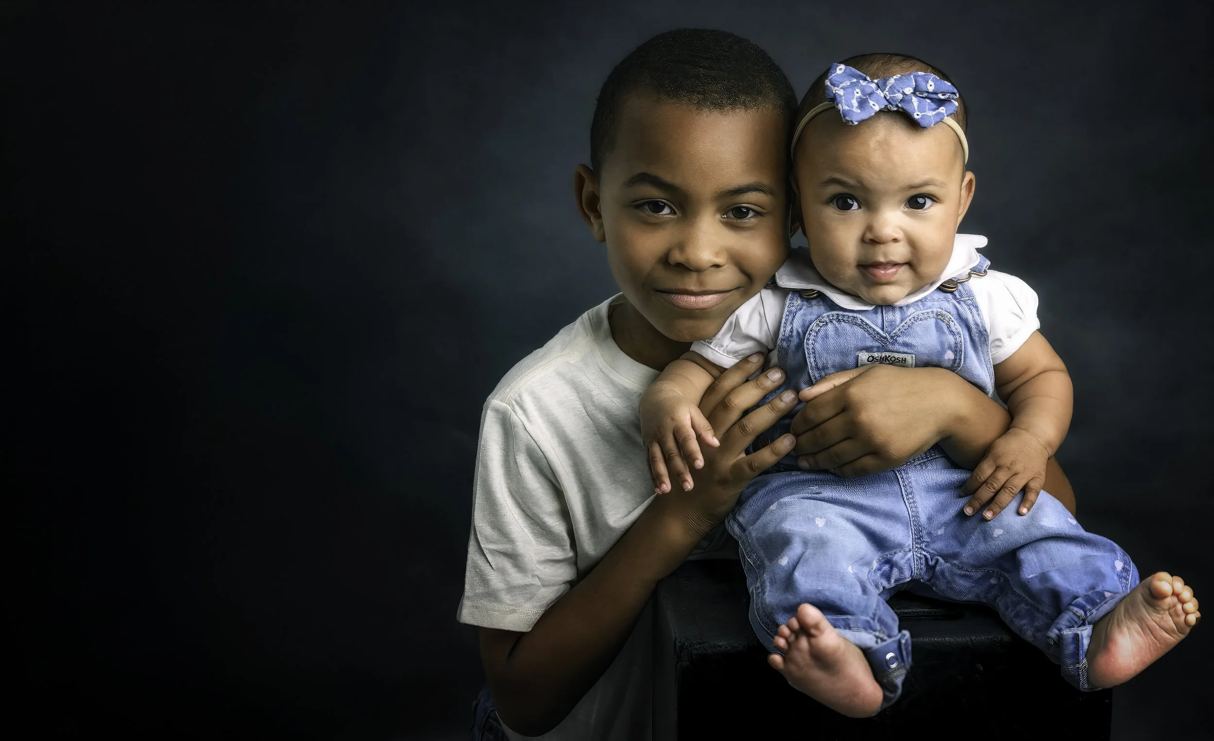 A young boy holding a baby girl against a dark background. The boy has short dark hair and wears a light-colored t-shirt. The baby girl wears blue overalls, a white shirt, and a headband with a blue bow. Both are smiling softly.