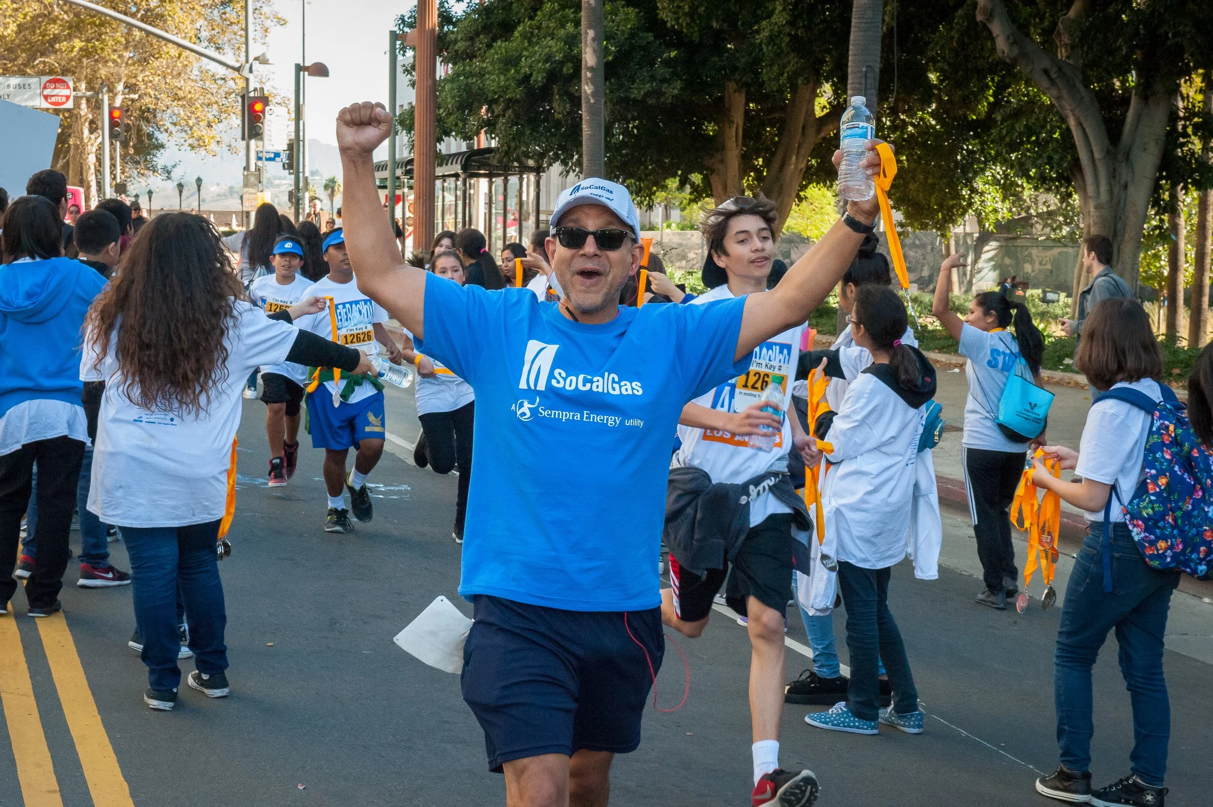 A man in sunglasses running in a race, raising his fist in celebration, surrounded by a group of children and teenagers at a marathon event, with trees and streetlights in the background.