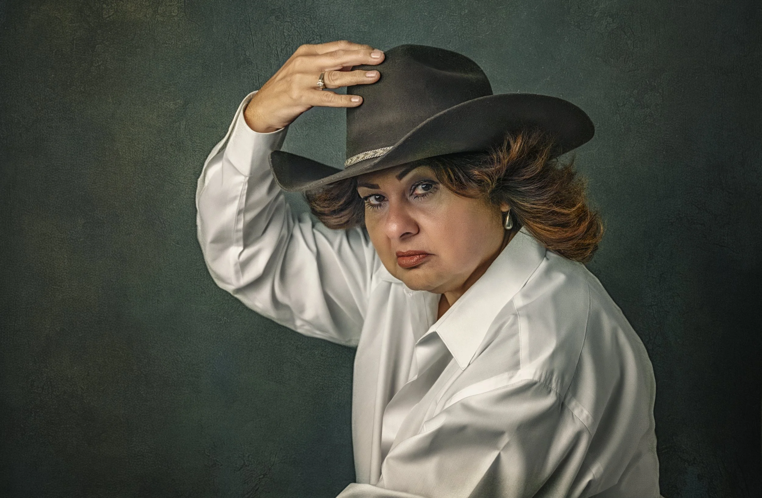 Creative portrait of a woman wearing a black cowboy hat and white shirt against a textured green background.