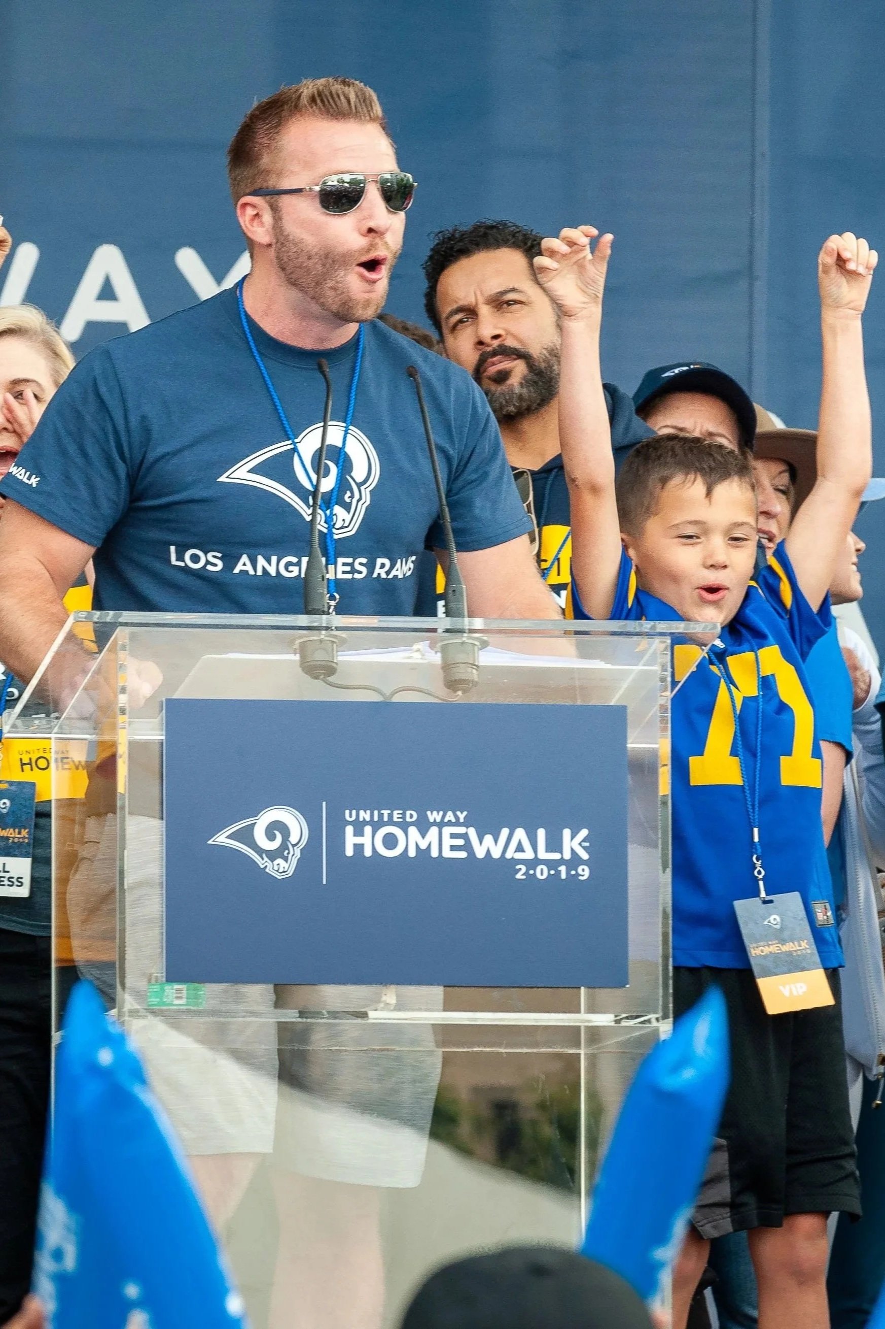 Sean McVay, wearing sunglasses, a blue Los Angeles Rams t-shirt, and a blue wristband is speaking at a podium during a United Way Homewalk event in 2019. Behind him, a diverse group of people