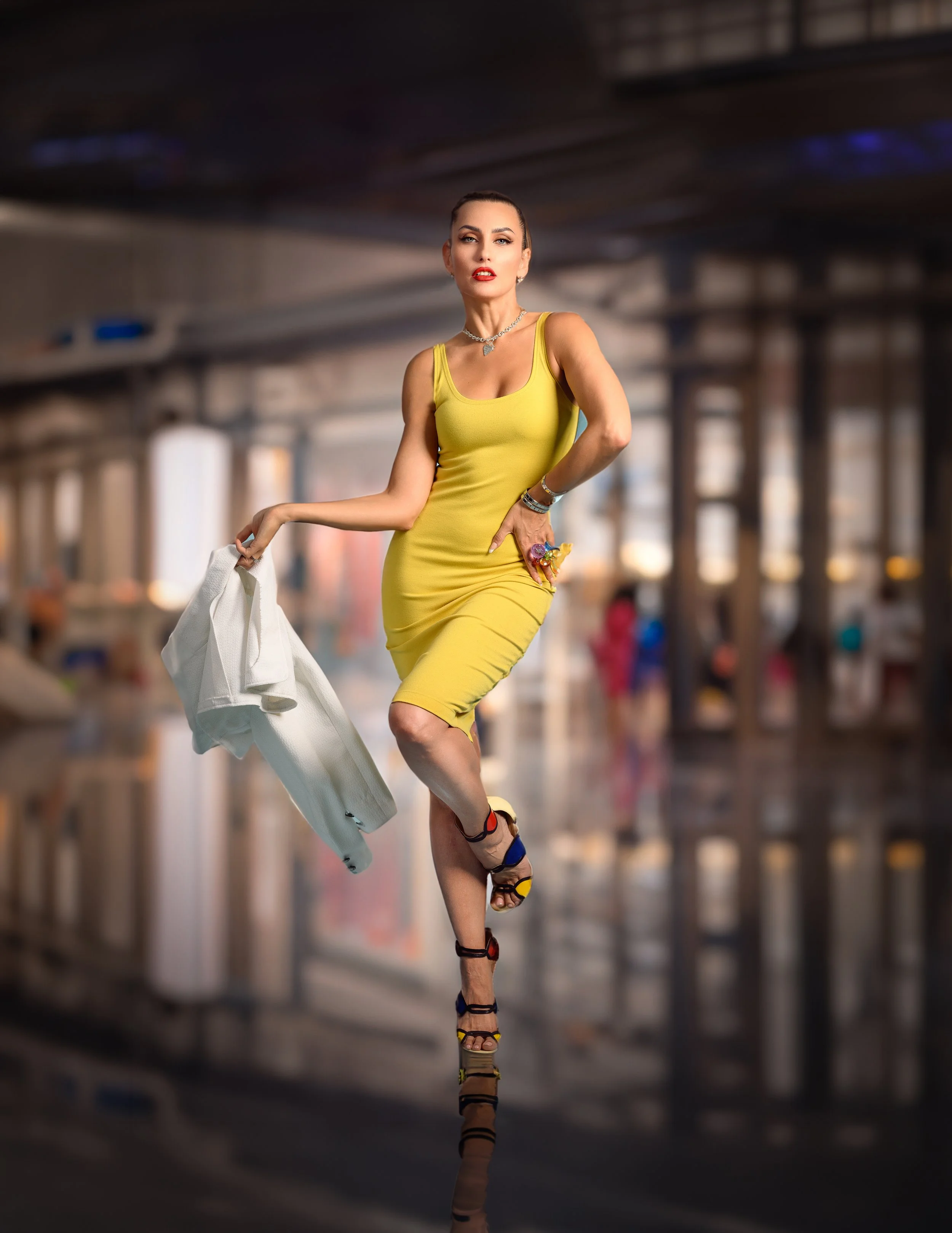 A woman in a yellow dress walking in an airport or shopping mall, holding a white jacket, with blurred figures and structures in the background.
