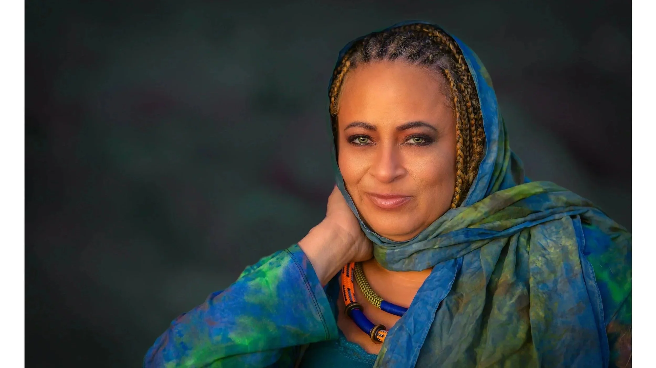 A woman with braided hair wearing a colorful scarf around her head and a tie-dye jacket, looking at the camera with a slight smile against a dark background.