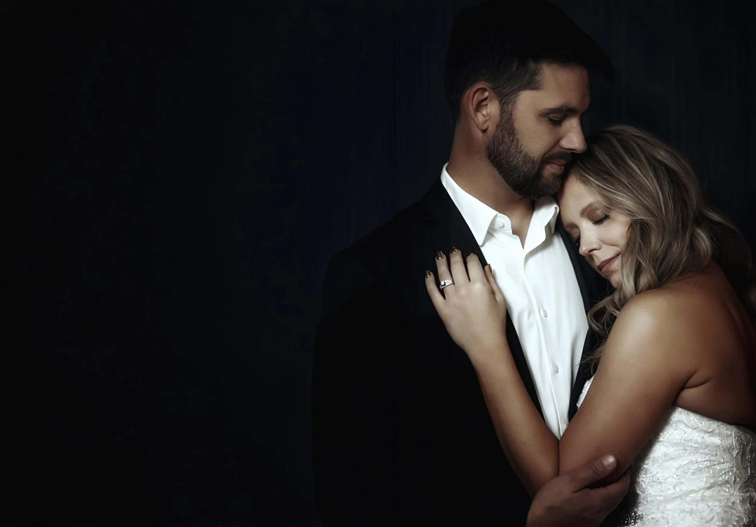 A groom and bride embracing closely, with their eyes closed, against a dark background.
