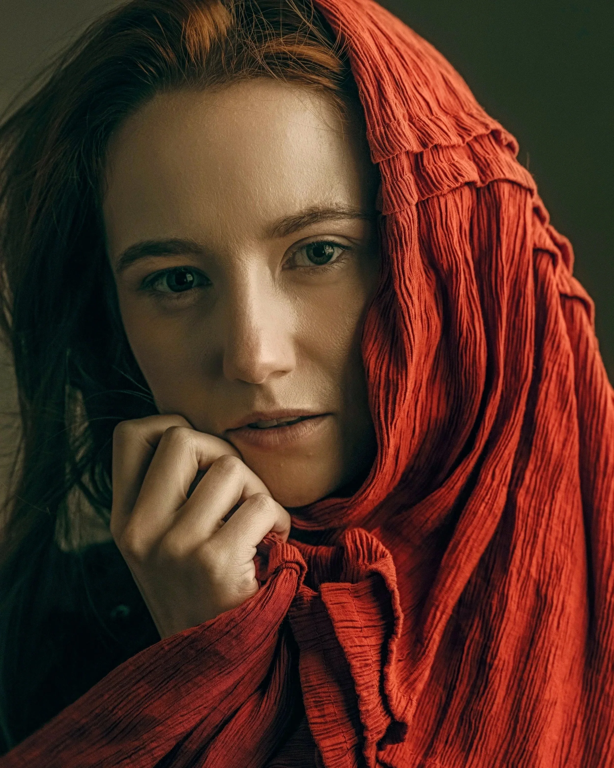 A woman with long brown hair, light skin, and striking blue eyes, wearing a red textured scarf or shroud that covers part of her face and shoulder, with a contemplative expression, set against a neutral dark background.