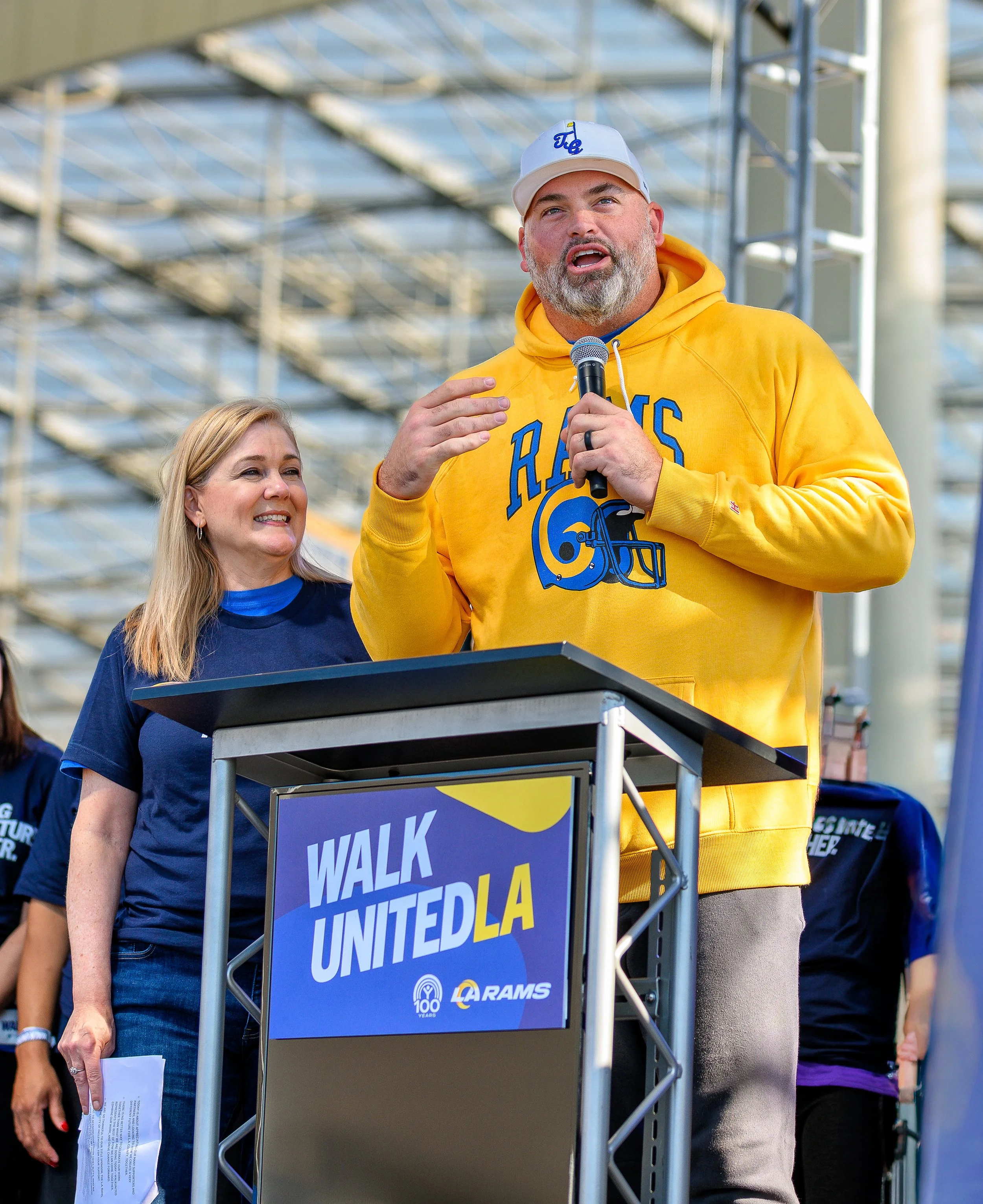 A man with a beard wearing a yellow hoodie with a sports logo, holding a microphone and speaking at a podium during an outdoor event, with a woman standing beside him. The background includes a metal structure and other people.
