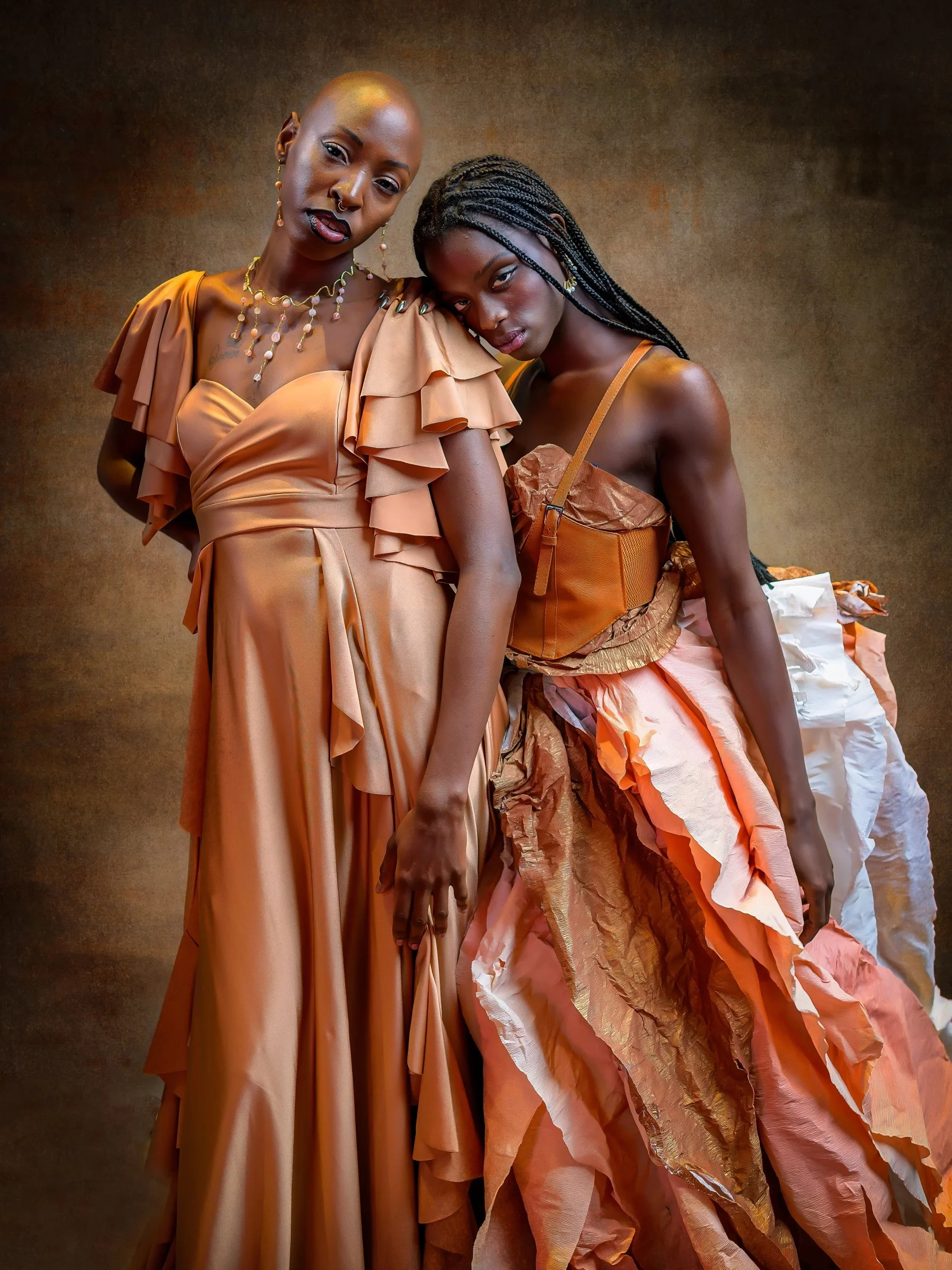 Two women in elaborate fashion with gold and peach-colored outfits. One woman has a shaved head, tattoos, and jewelry, while the other has long braided hair and a ruffled, textured skirt. They are posed closely against a textured brown background.