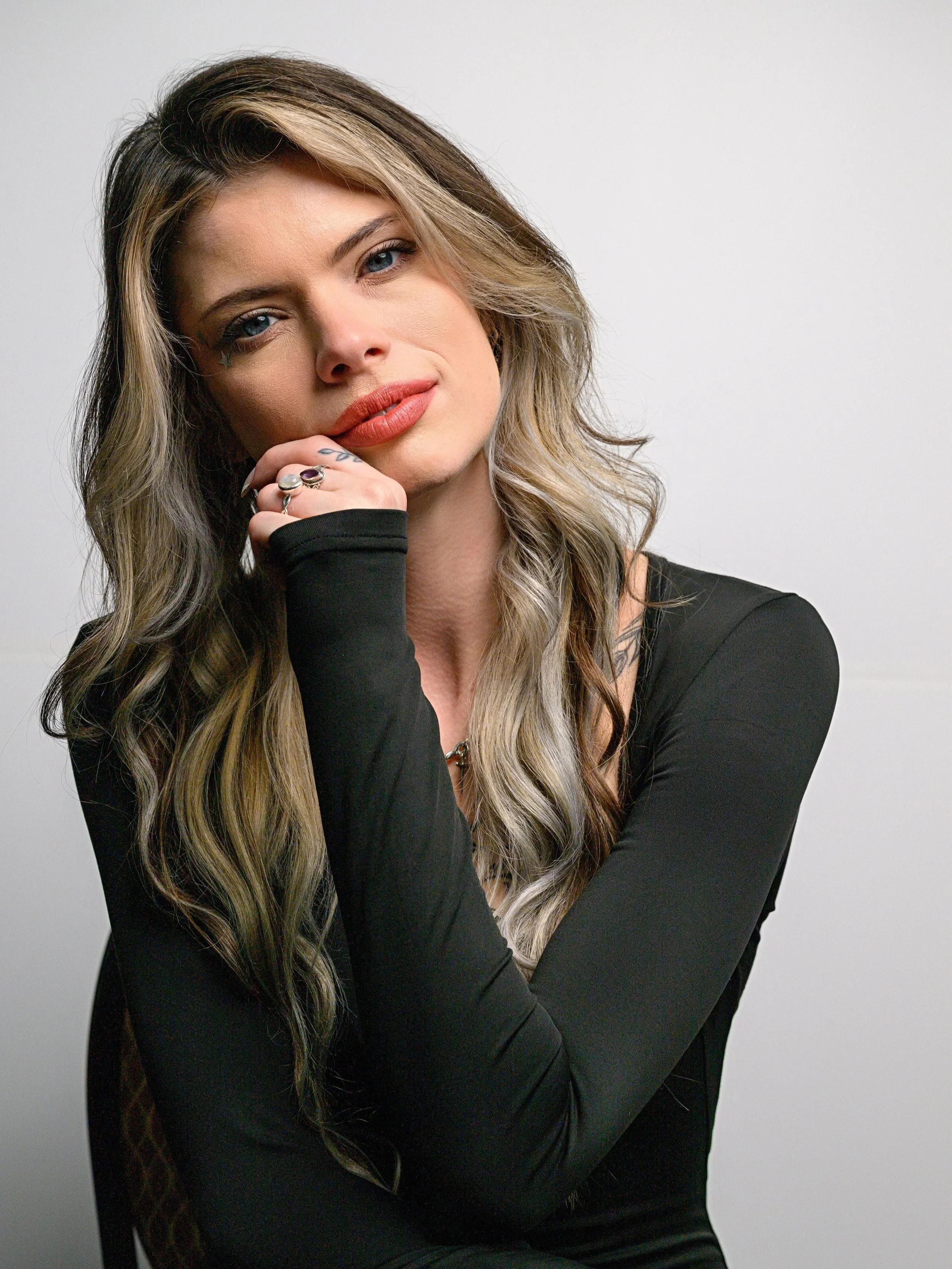 A woman with long, wavy hair and blue eyes, wearing a black long-sleeve top, with a small star tattoo under her right eye, rings on her fingers, and a confident expression, posing against a plain light-colored background.