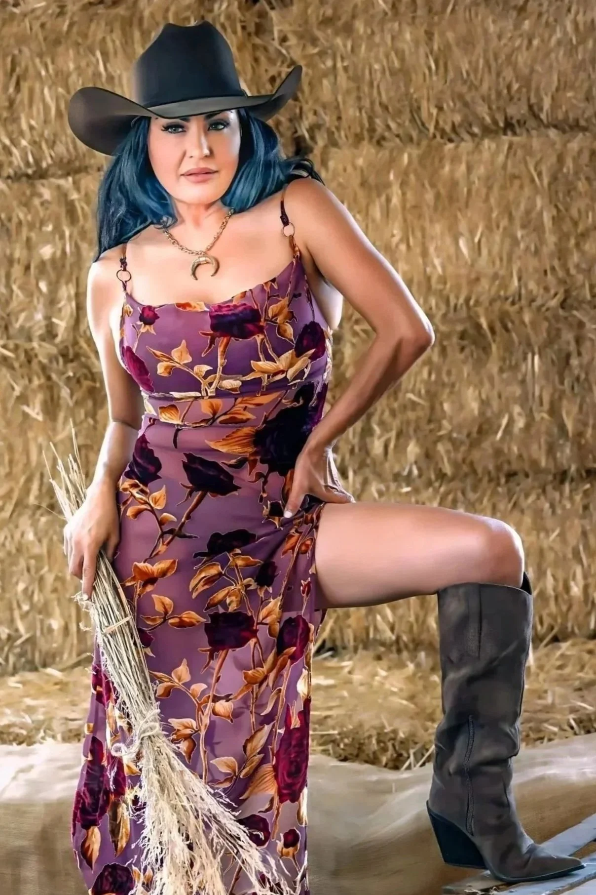 A woman with black hair wearing a wide-brimmed cowboy hat and a floral dress with slits, holding a bunch of dried grass, standing in front of stacked hay bales, with a hand on her hip, and wearing black cowboy boots.