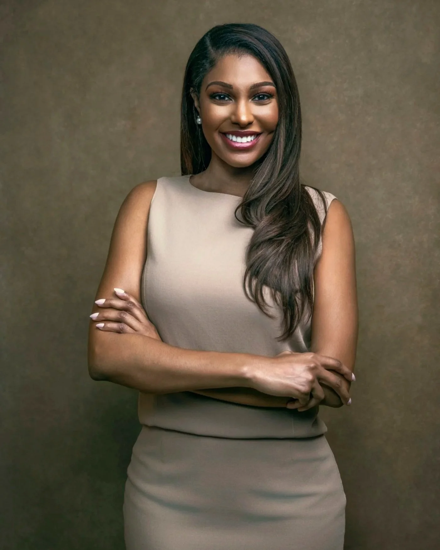 A woman with long dark hair wearing a sleeveless beige dress, smiling with arms crossed against a neutral background.