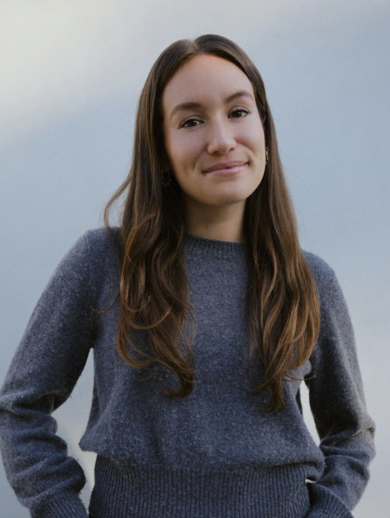 A young woman with long brown hair, wearing a dark gray sweater, standing outdoors against a cloudy sky.