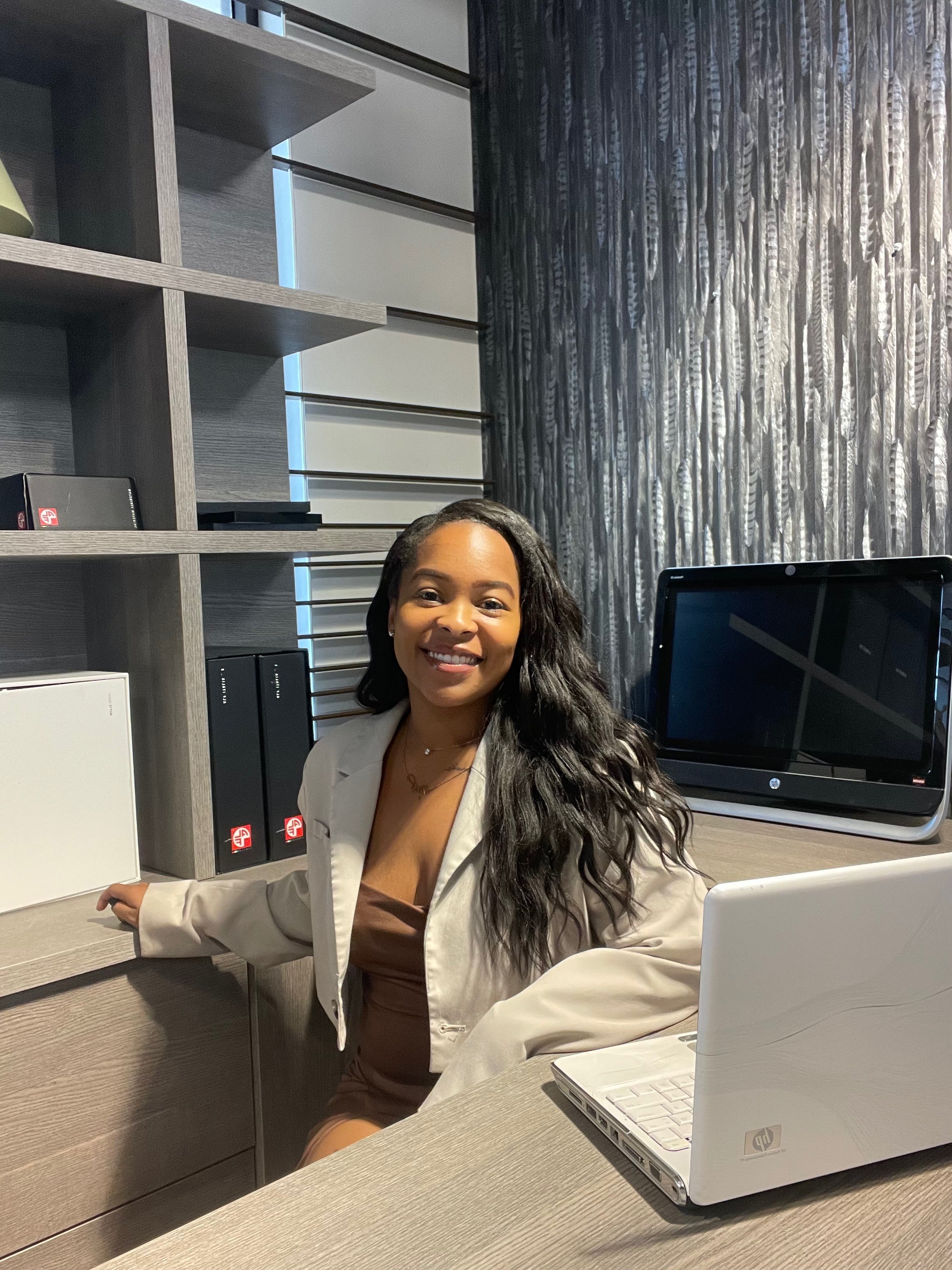 A woman with long, dark, wavy hair and a bright smile sitting at a desk in an office, wearing a beige blazer and brown dress, with a white HP laptop on the desk, behind her a gray wall with shelves and a window with patterned gray curtains.