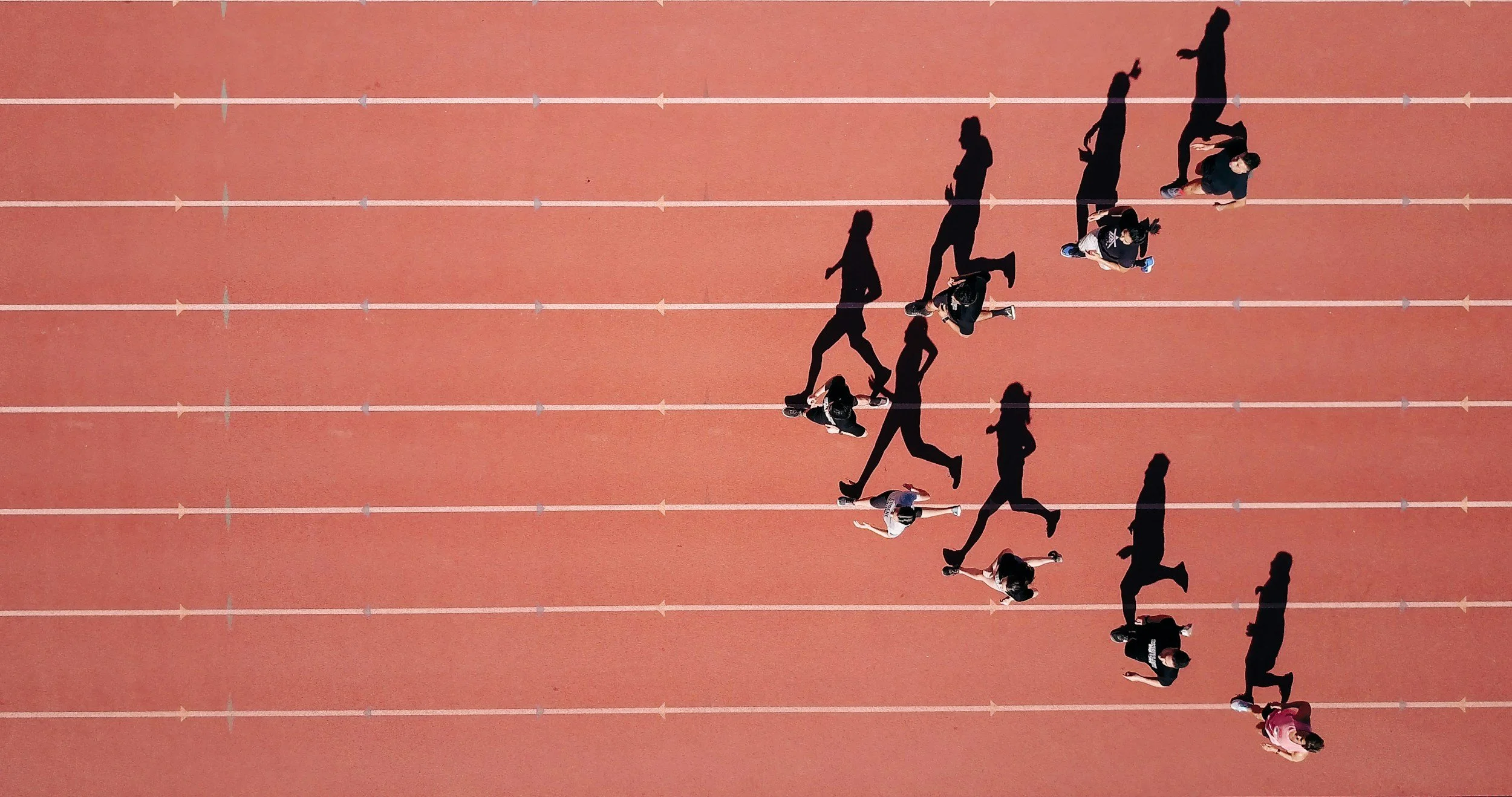 People running on a red outdoor athletic track, seen from above, casting long shadows.