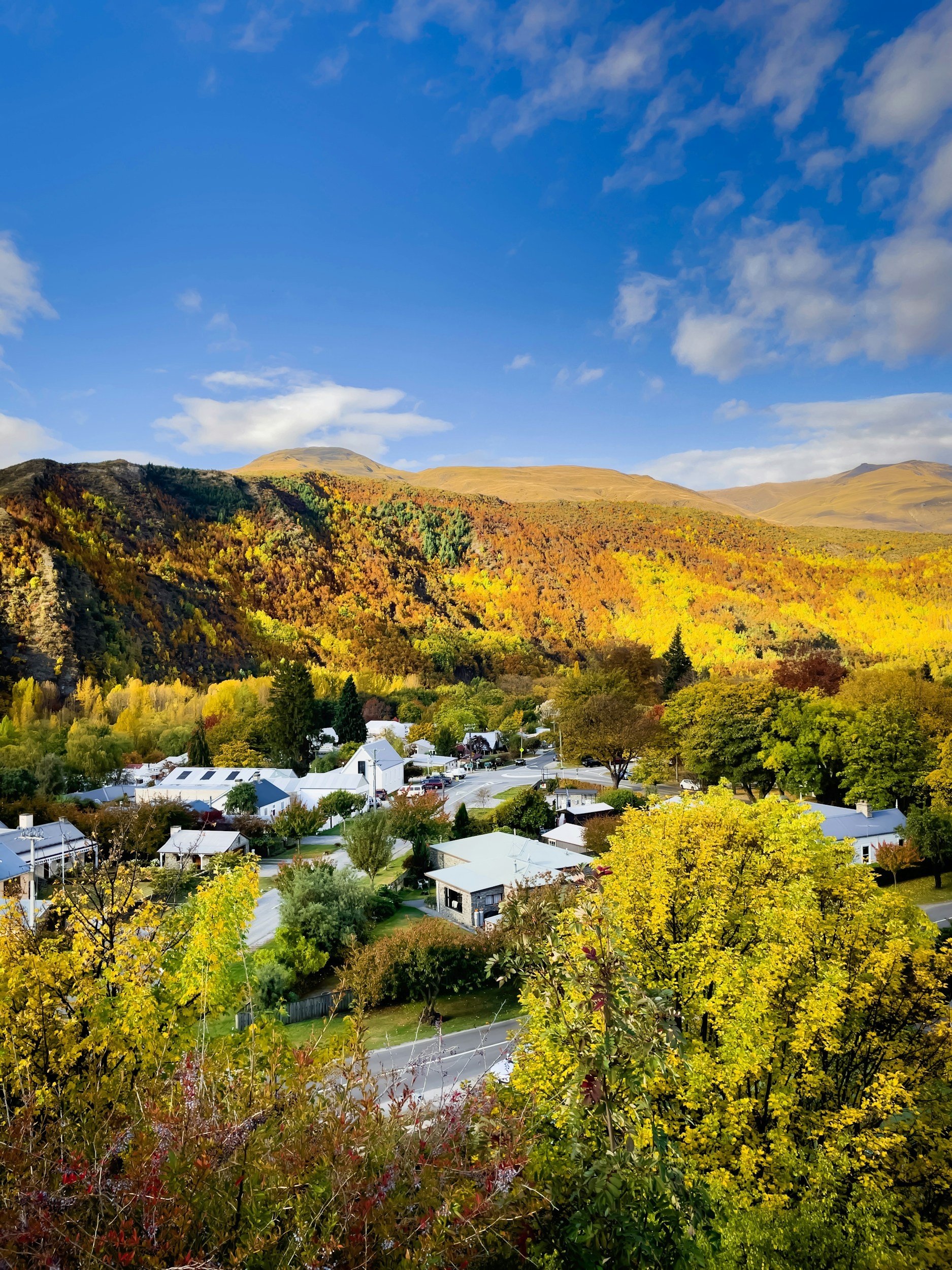 Scenic view of a small town surrounded by colorful autumn trees, hills, and mountains under a partly cloudy blue sky.