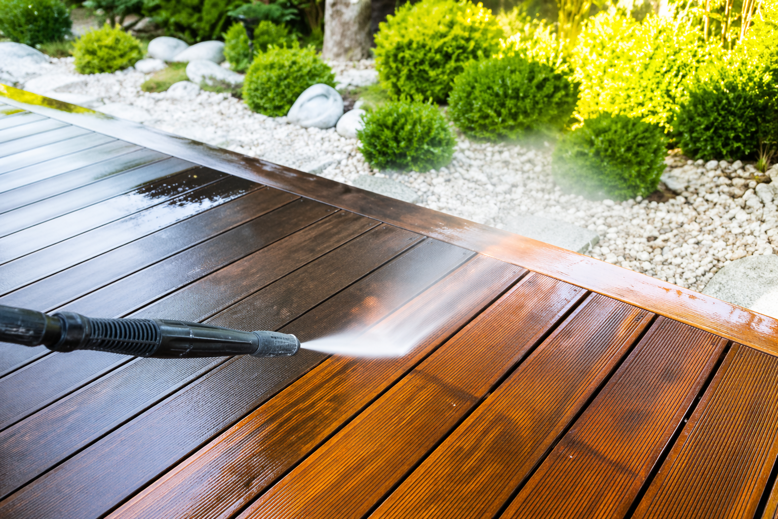A pressure washer cleaning a wooden deck outdoor area with plants and rocks in the background.