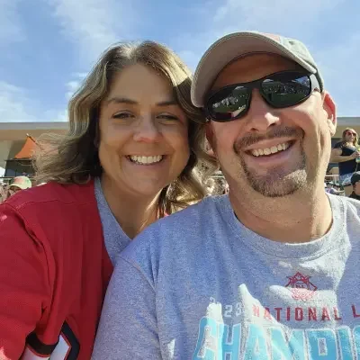 A smiling man and woman taking a selfie outdoors on a sunny day, with a blue sky and a volleyball net in the background.