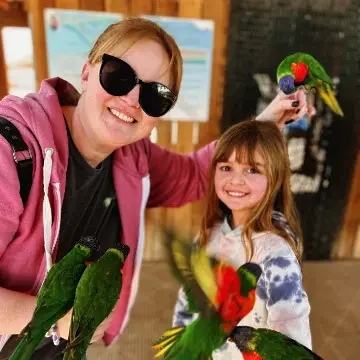 A woman and a girl holding colorful parrots, smiling indoors with a blurred background.