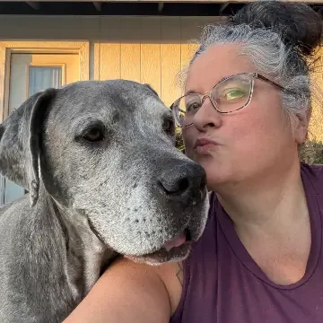 A woman with gray hair pulled back, wearing glasses and a purple shirt, taking a close-up selfie with a large gray and white speckled dog.