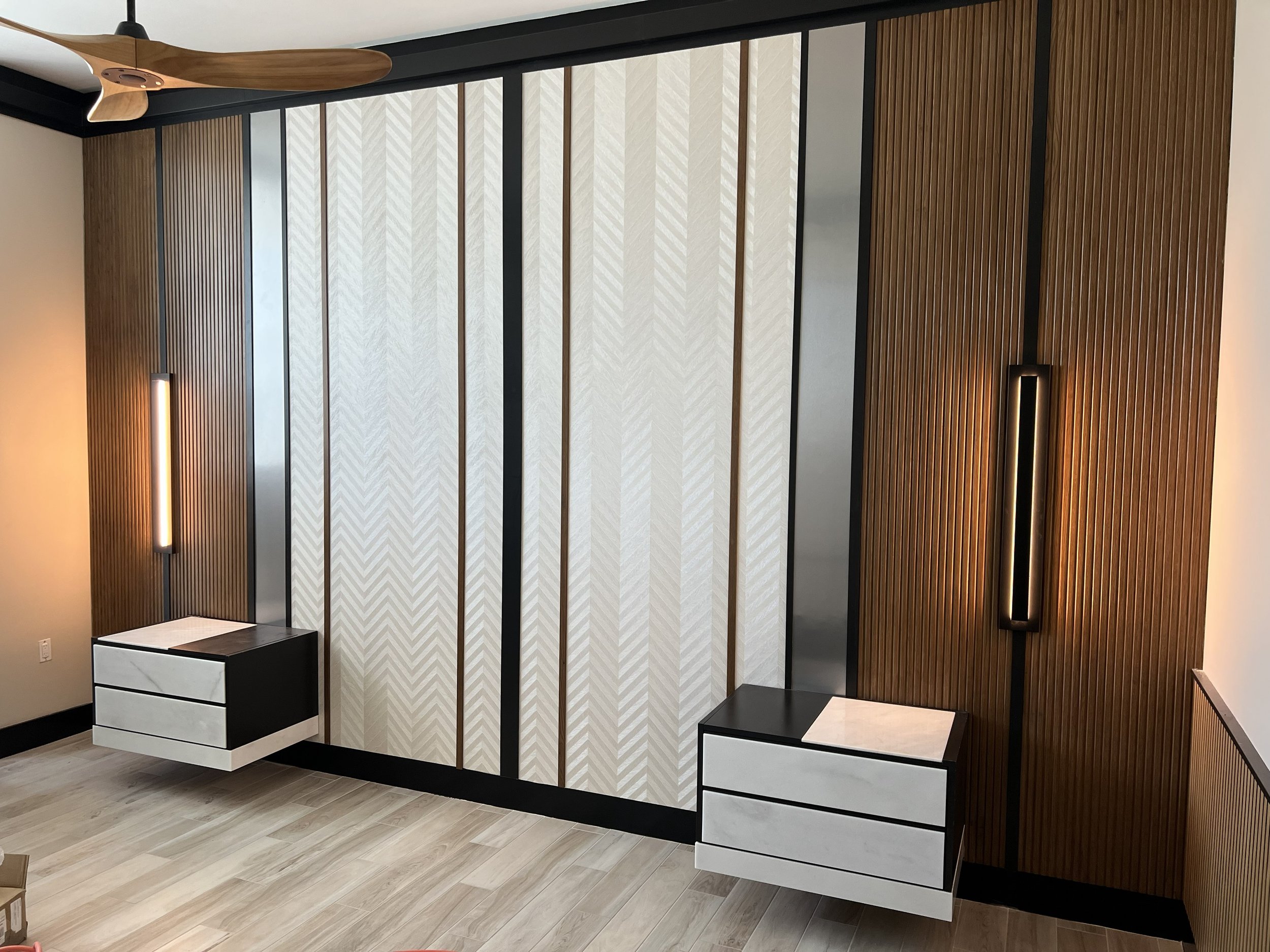 Modern decorative wall with wood slats and patterned panels, black metal accents, and built-in lighting, with white and black night stands.