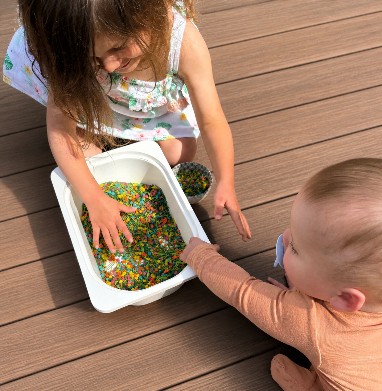 Two children, a girl and a baby, sitting on a wooden deck, playing with colorful rainbow sprinkles from a white container, with a small bowl of sprinkles nearby.