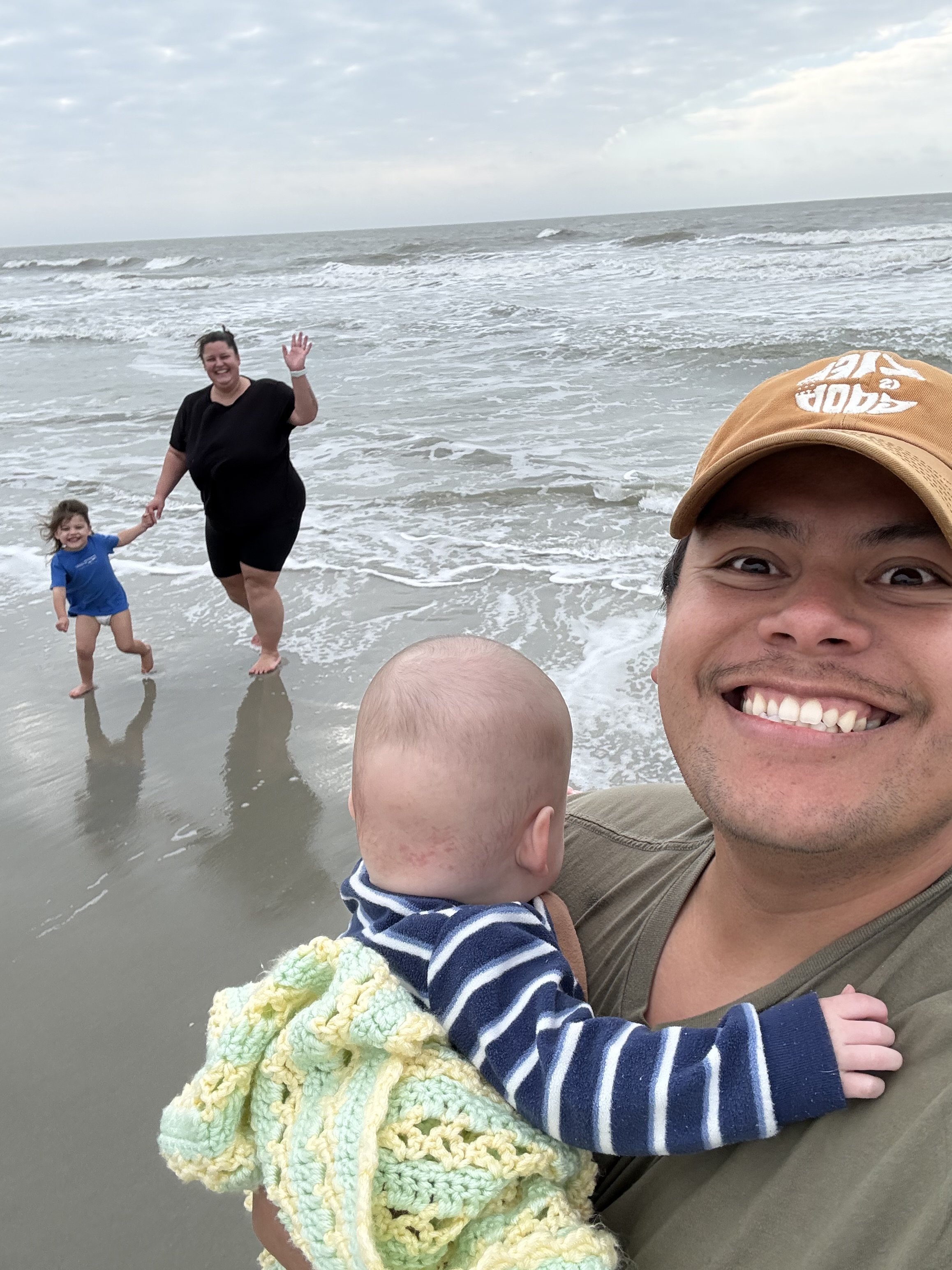 A family enjoying time at the beach, with a smiling man taking a selfie, a woman and child playing in the water, and the ocean in the background.