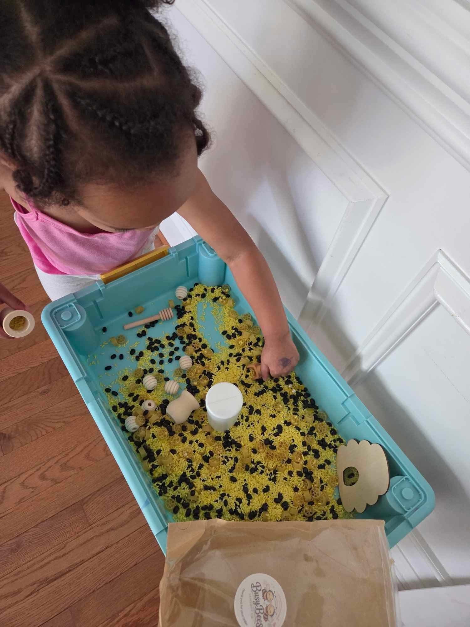 A young girl with braided hair playing with yellow rice and black beans in a bee themed sensory kit. She is promoting how engaging the Signature Kit is for Busy Bees Sensory Co.