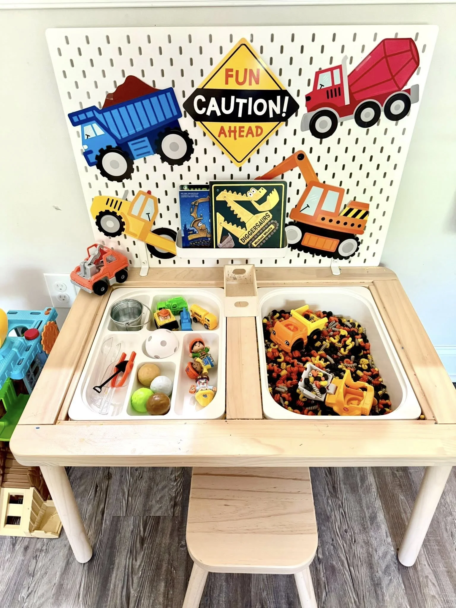 Wooden play table with two bins filled with small toy vehicles and other learning tools. Attached to the back of the table is a hanging display with construction vehicle images and a caution sign.