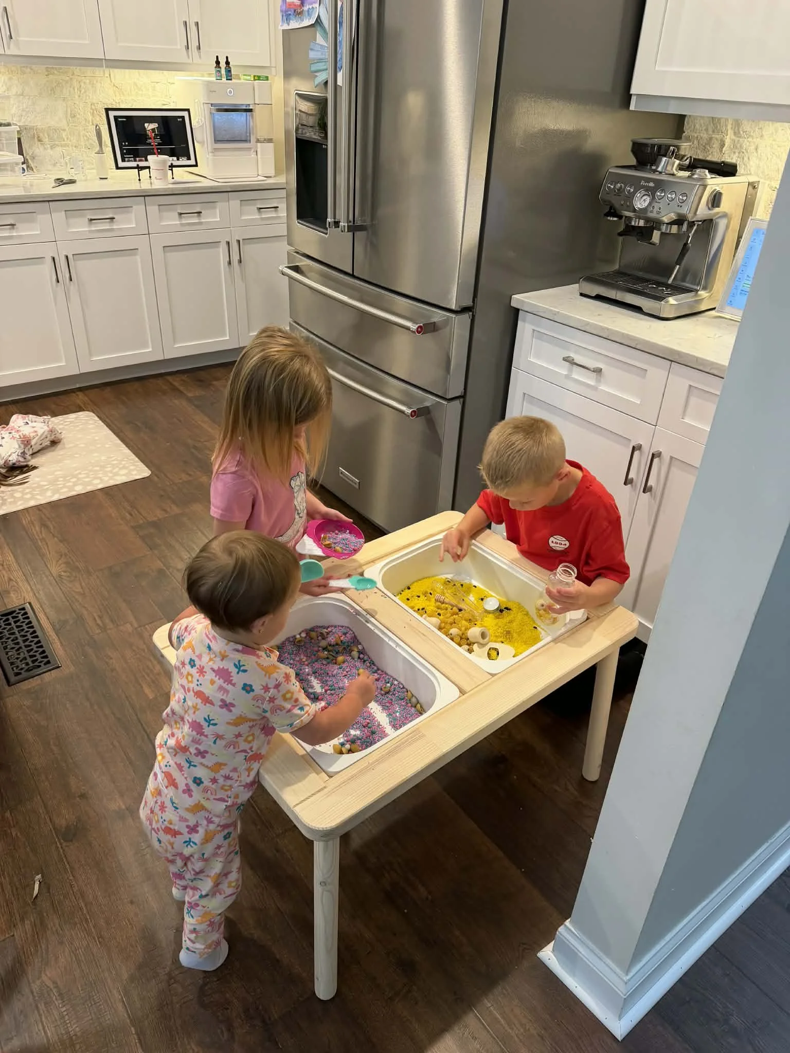 Children playing with sensory bins filled with colorful beads, rice, and other items in a kitchen. Two kids are standing and looking into the bins, engaging with their surroundings.