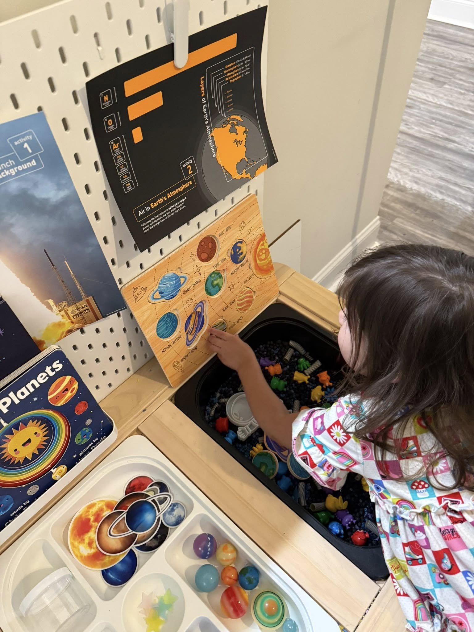 A young girl wearing a colorful pajama set is reaching into a box filled with marbles of various colors and sizes. There are educational posters about planetary science and Earth's atmosphere displayed on a pegboard behind her.