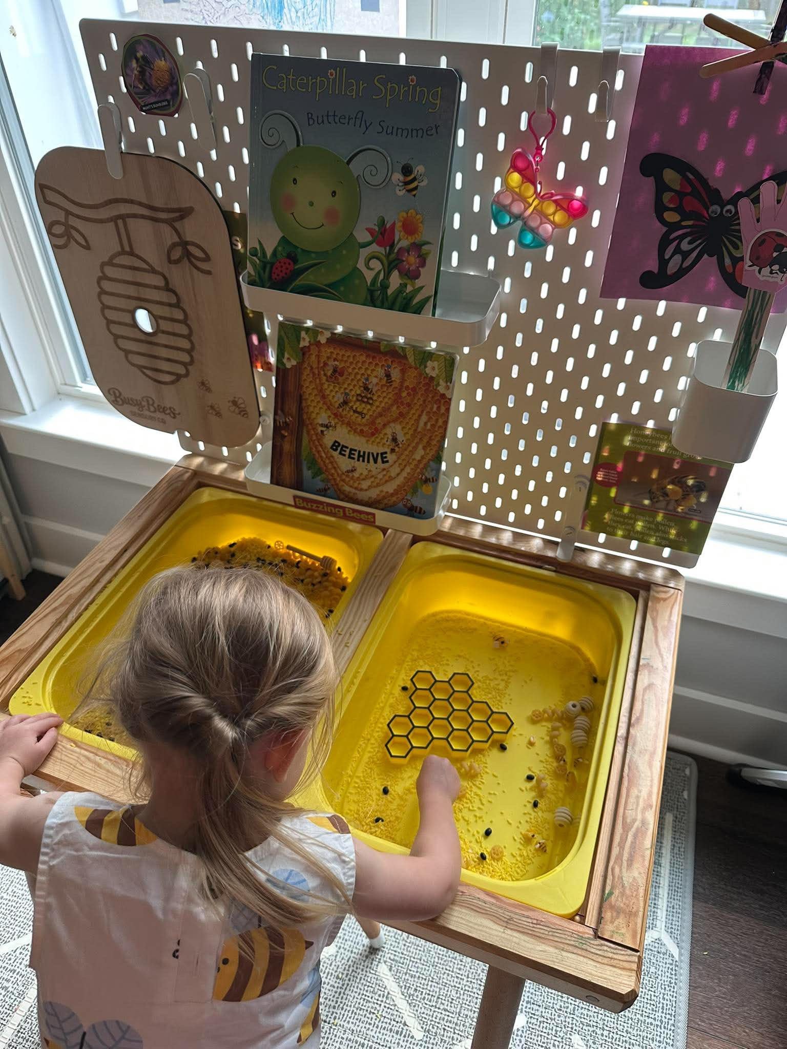 A young girl with blonde hair in pigtails playing with a yellow honeycomb-themed sensory bin filled with plastic bees, black beads, and honey bee-themed toys, at a wooden table in a bright room.