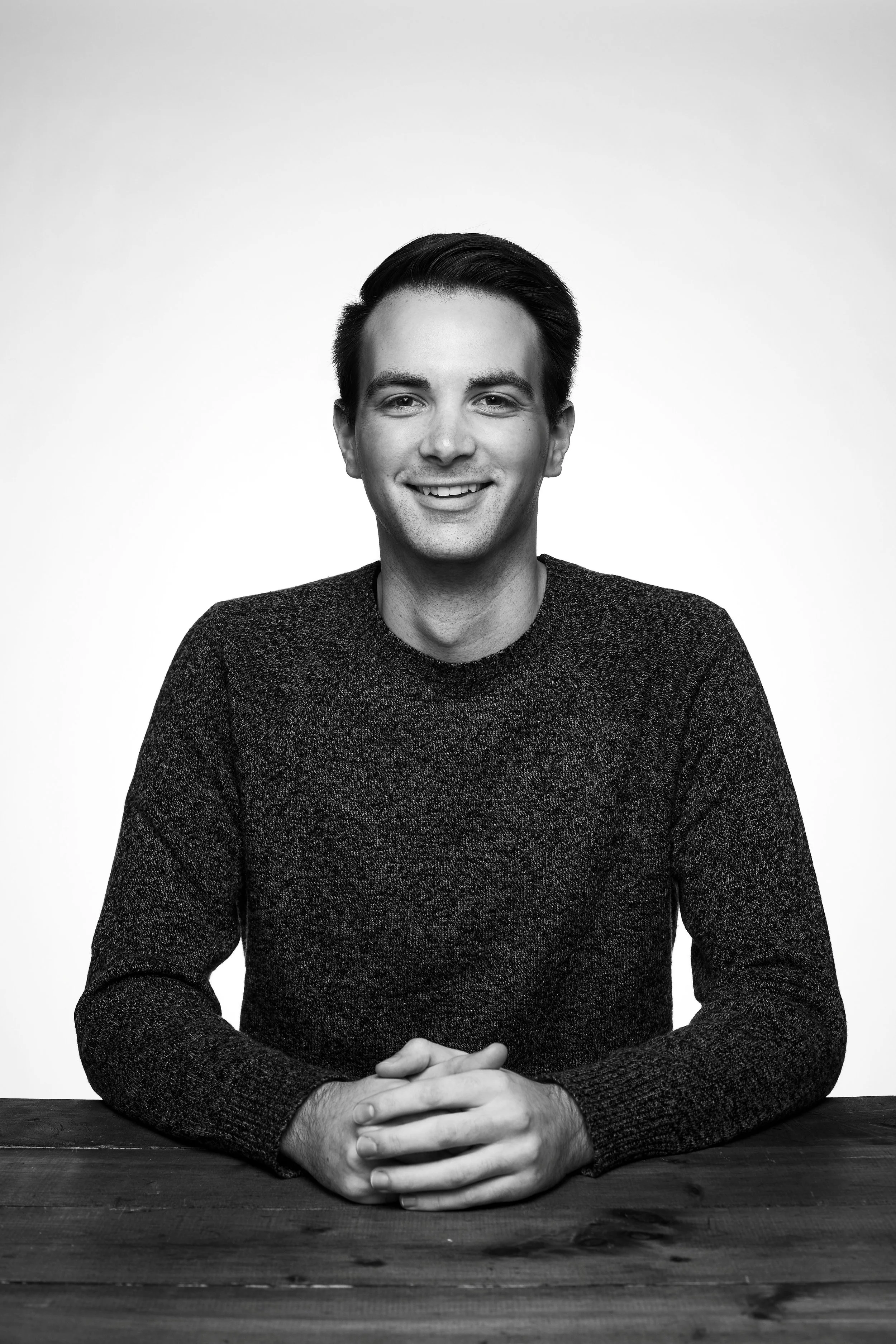 Black and white portrait of a young man with dark hair, smiling, wearing a dark sweater, sitting at a table with hands clasped on the surface.