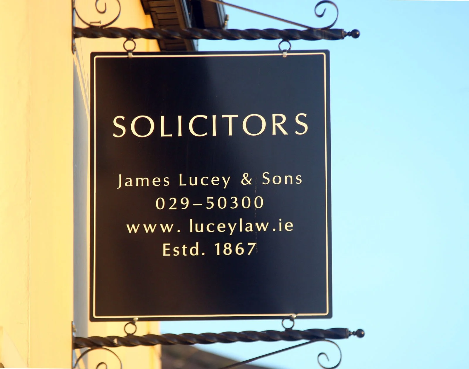 Black and white signboard for solicitors, showing the name James Lucey & Sons, contact number 029-50300, website www.lucy law.ie, established in 1867, hanging from a decorative wrought iron frame.