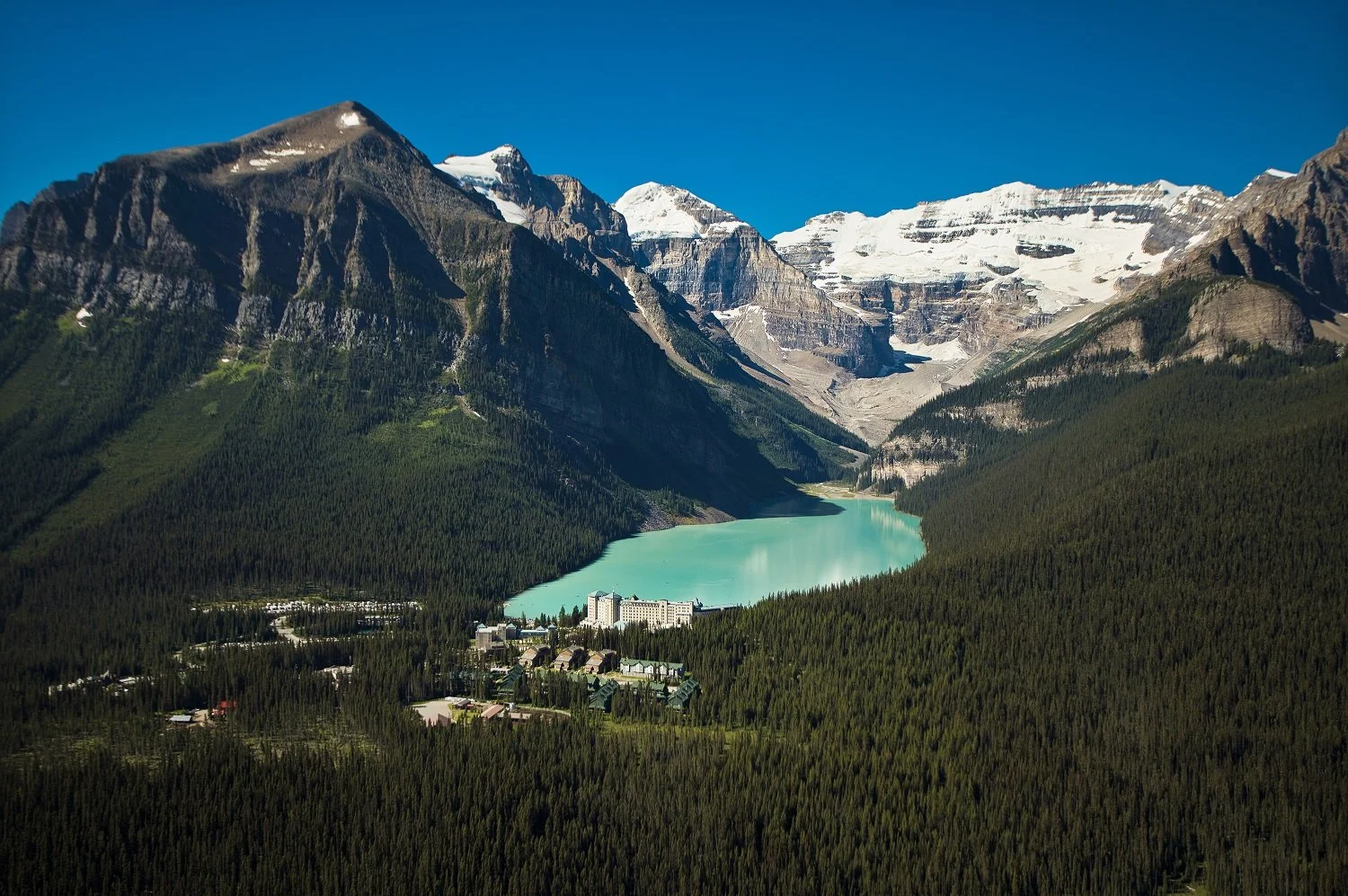 Mountain landscape with snow-capped peaks, a turquoise lake, and a forested valley with buildings in the foreground.