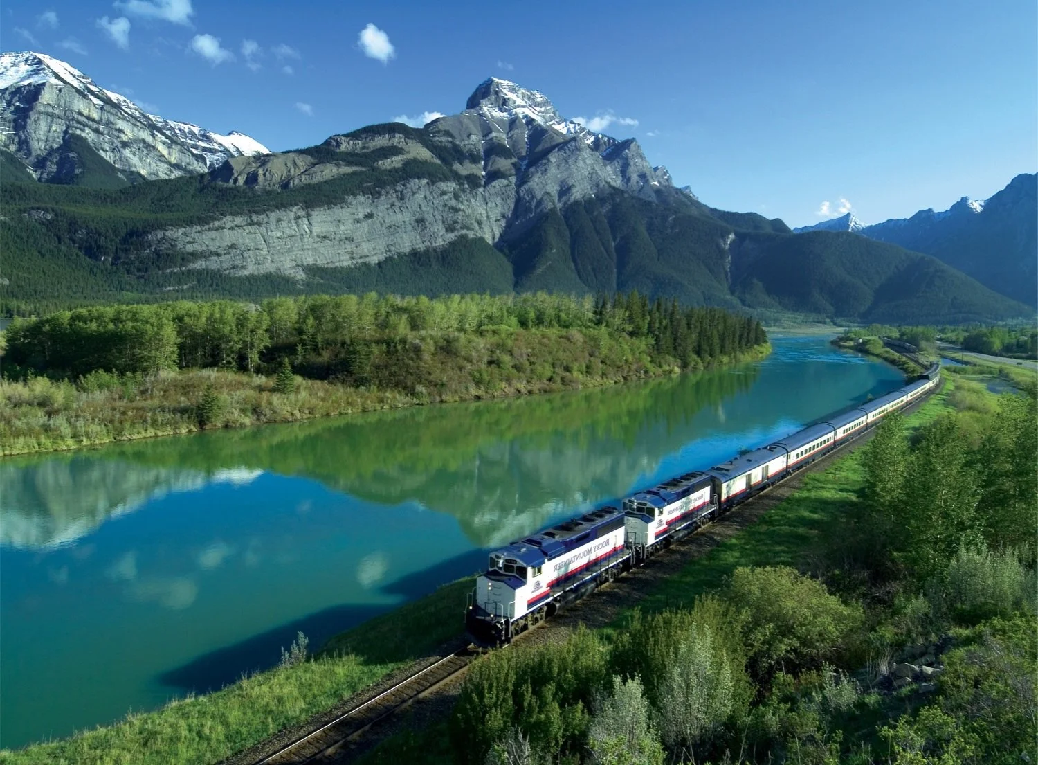 A scenic landscape with a train running alongside a river with mountains in the background, lush green trees and a bright blue sky.