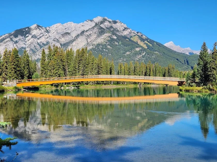 A scenic view of a mountain range, dense forest, a calm river with a reflection, and a yellow bridge under a clear blue sky.
