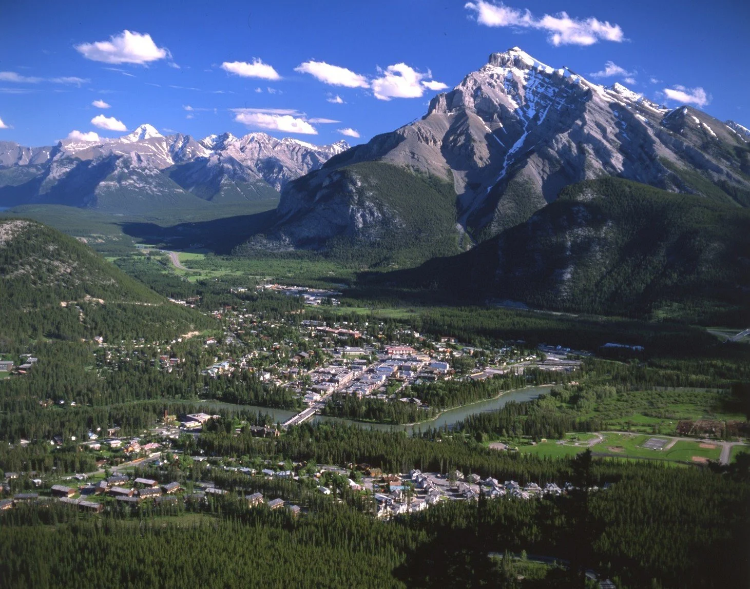 Aerial view of a town nestled in a green valley with large mountains, some with snow caps, in the background under a blue sky with scattered clouds.