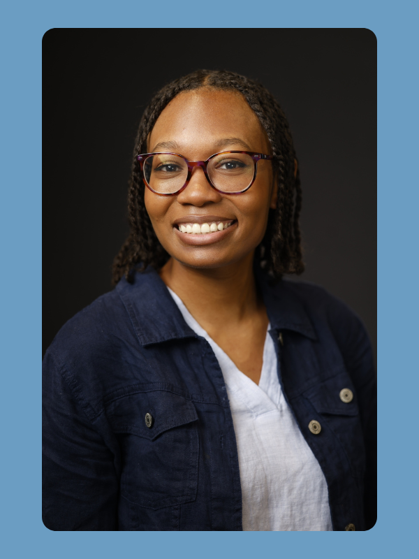 A smiling woman with glasses and braids wearing a white shirt and dark blue jacket against a dark background.