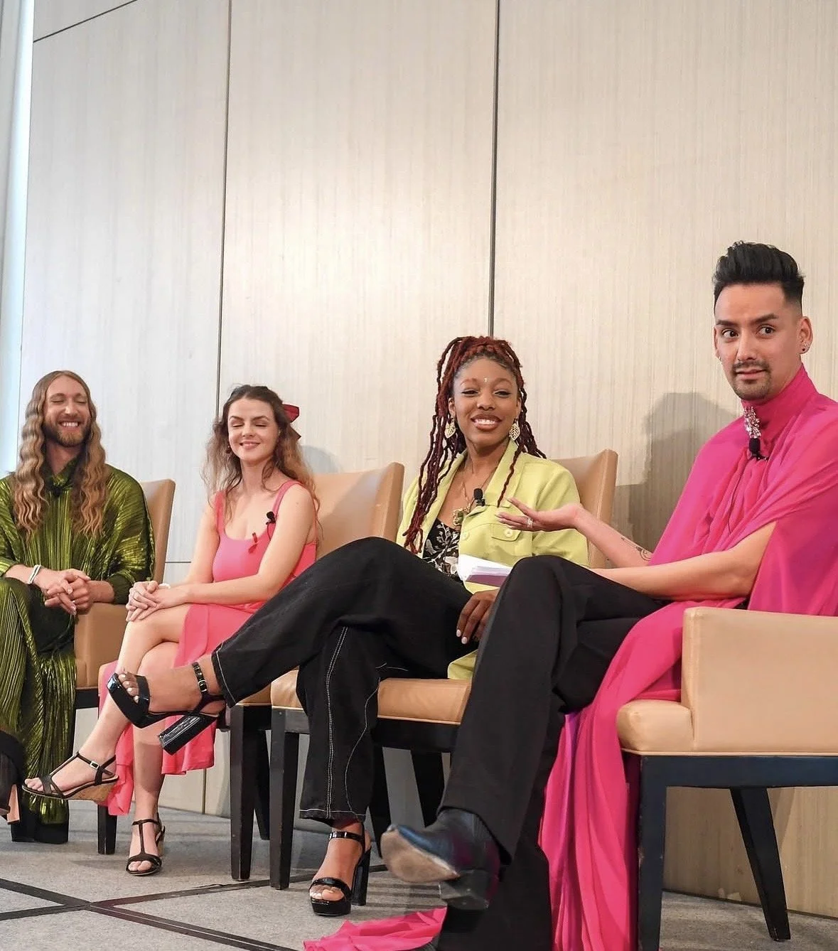 Four diverse individuals sitting and smiling on a panel at an event, dressed in colorful attire, with a plain wall background.
