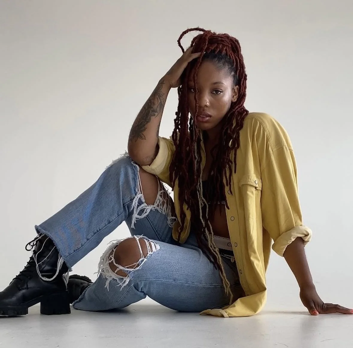 Tyra Blizzard, professional basketball player, Global Partnerships & Inclusive Marketing Consultant, sitting on the floor, wearing a yellow shirt, ripped jeans, and black boots, against a plain white background.