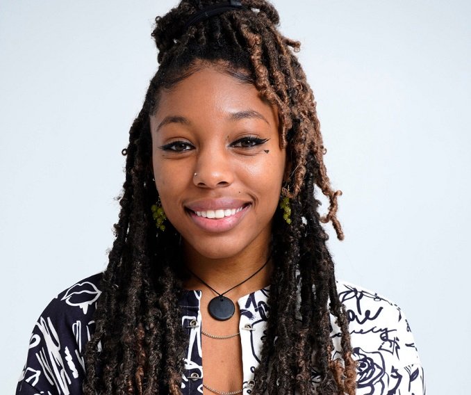 Young woman with dreadlocks, smiling, wearing a black and white patterned shirt, jewelry, and a hairband.