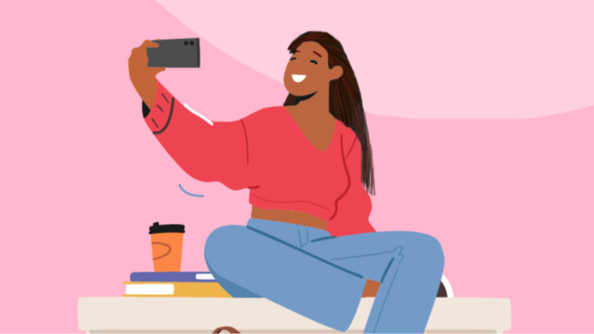 A woman sitting cross-legged on a desk, taking a selfie with her phone, with a coffee cup and books nearby, against a pink background.