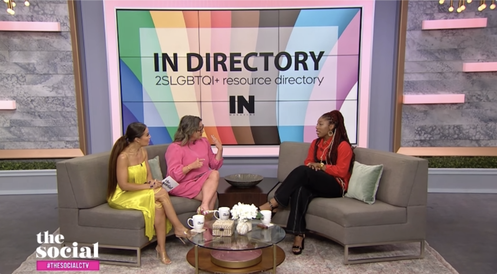 Three women participating in a talk show on a set with a large screen behind them displaying 'IN DIRECTORY' and '2SLGBTQI+ resource directory'. They are seated on couch chairs, with a coffee table in front of them holding mugs, a small gift box, and a flower arrangement.