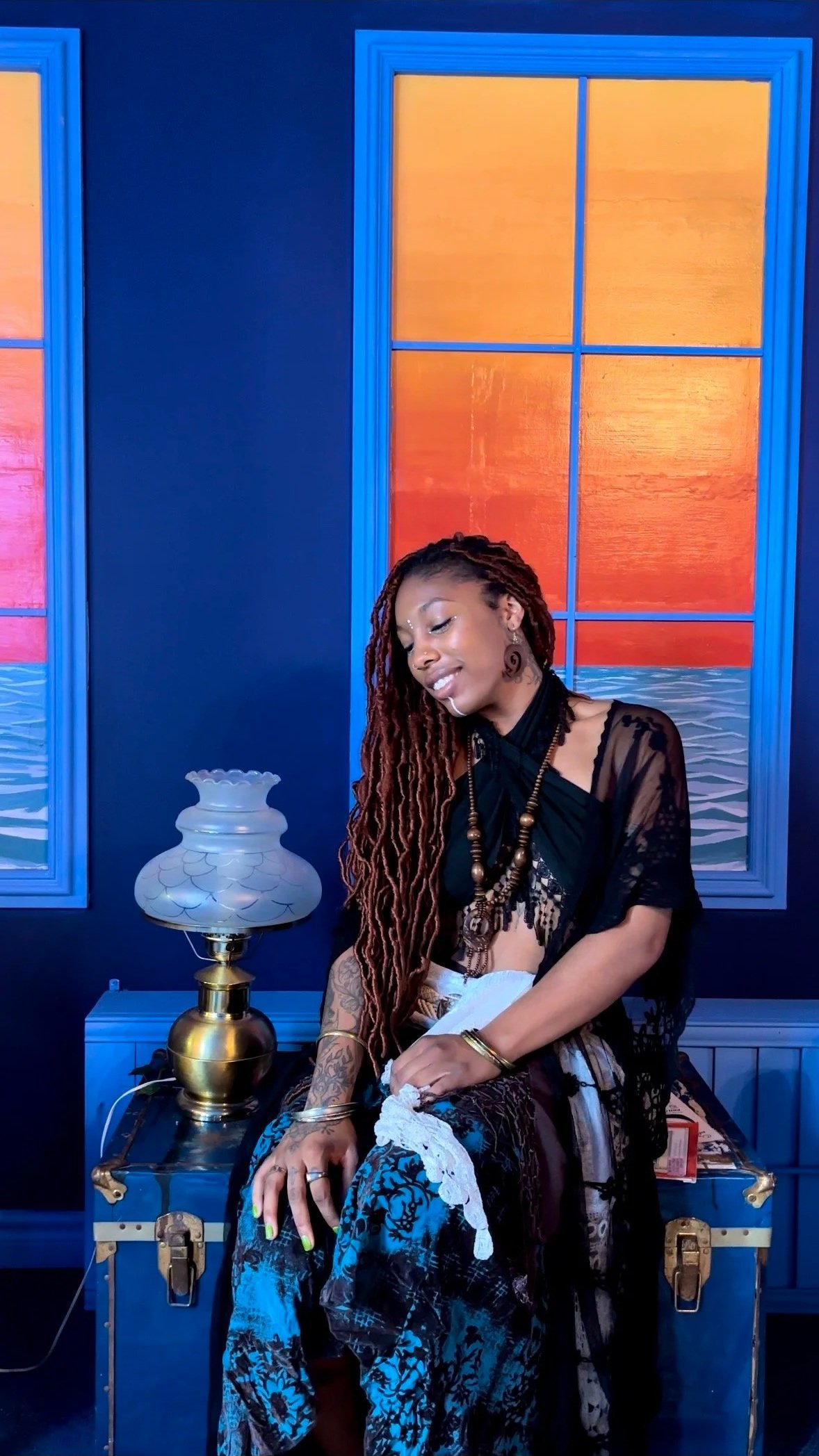 A woman with long braided hair, tattoos, and jewelry sitting on a blue chest in a colorful room with blue walls and orange window panels, next to a vintage lamp.