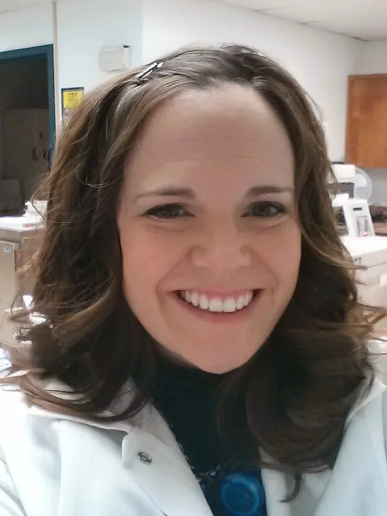 A woman with long wavy brown hair, wearing a white lab coat, smiling in a clinical or laboratory setting.