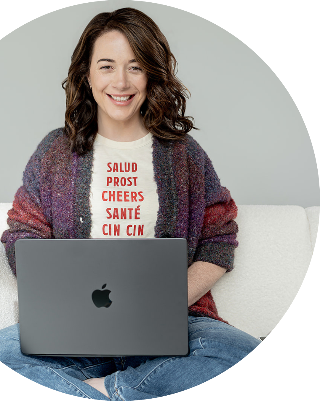 Brigitte Boots sitting cross-legged on a white couch, smiling at the camera with a silver MacBook on her lap, wearing a white T-shirt with red text and a colorful knitted cardigan.