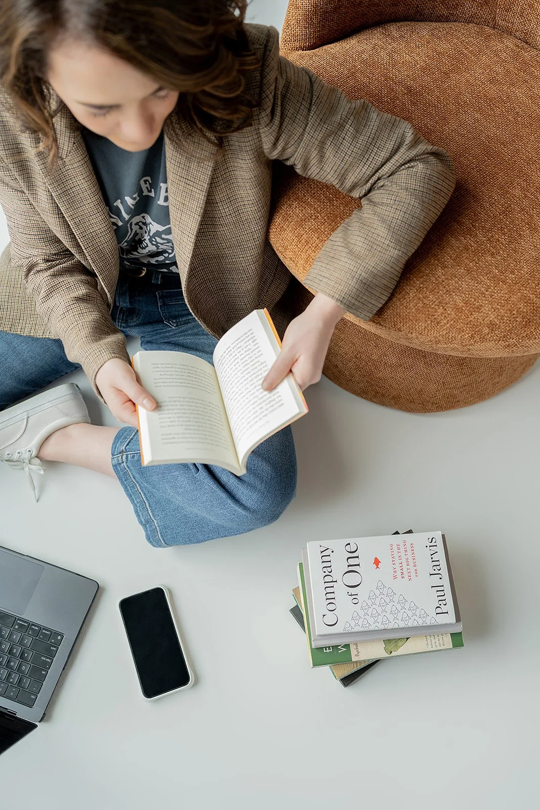 Brigitte Boots sitting cross-legged on the floor reading a book, with a laptop, a phone, and a stack of books on a table nearby, with an orange upholstered chair in the background.