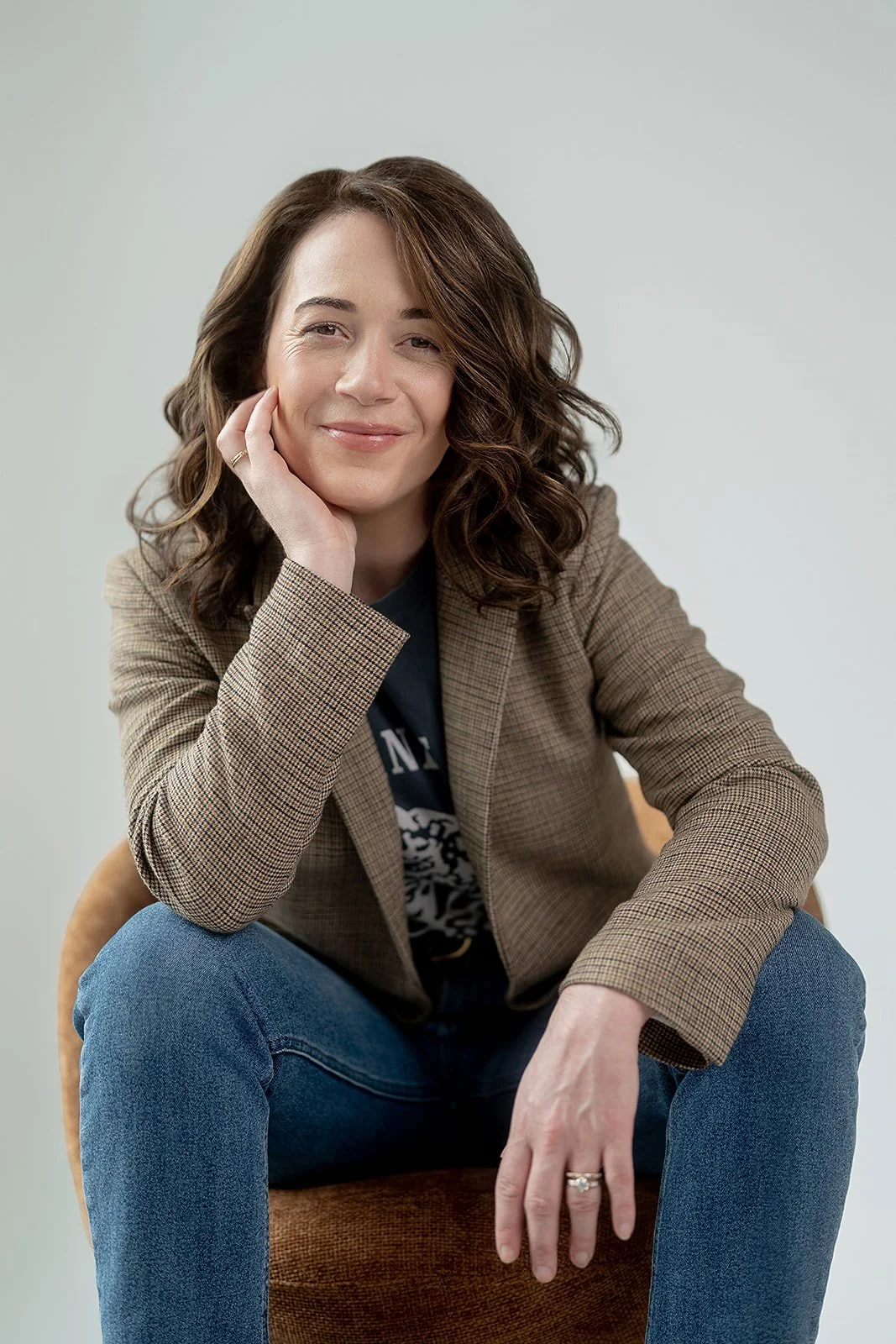 Brigitte Boots wearing a brown blazer and blue jeans, sitting on a chair against a plain light background, smiling softly and resting her chin on her right hand.