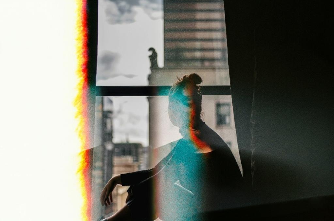 Silhouette of a person sitting by a window, with city buildings visible outside and a light leak effect on the left side of the photo.