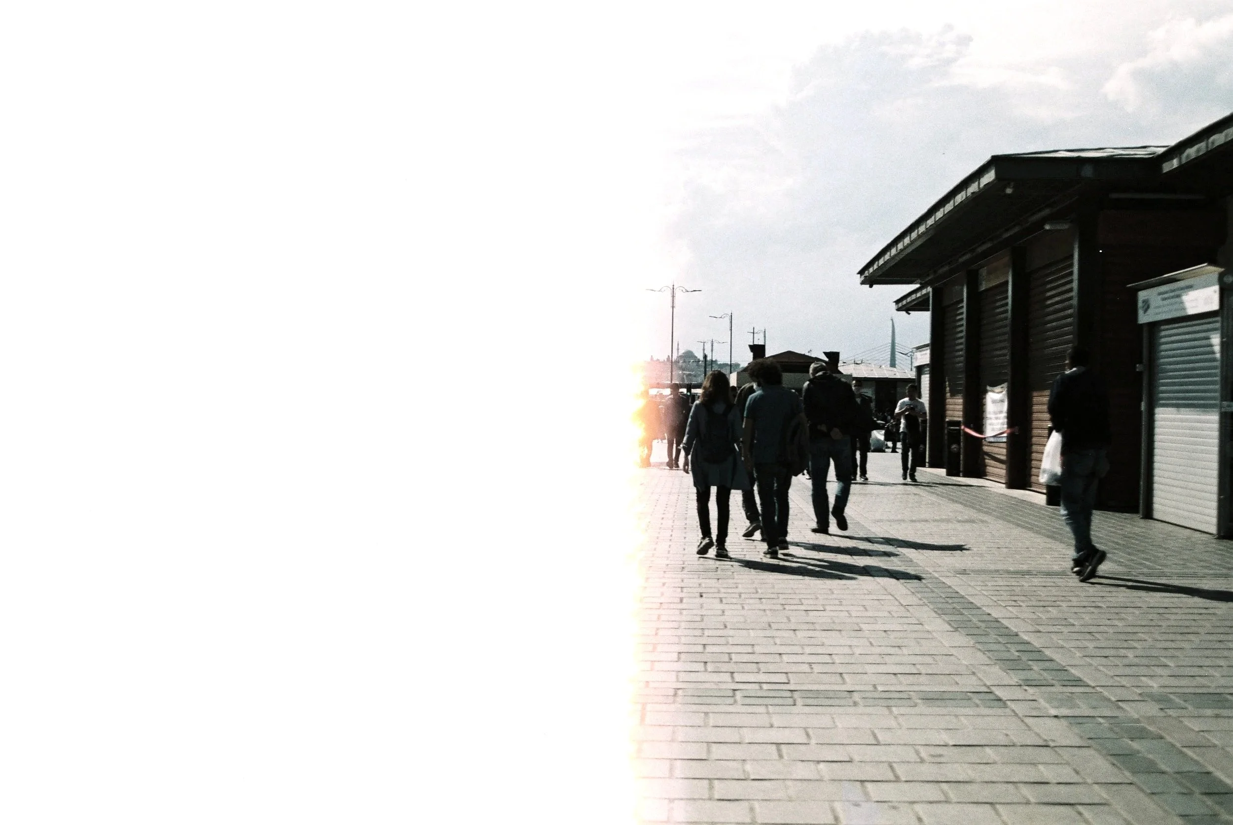 People walking on a sidewalk next to a row of closed shops with dark shutters, with a partly cloudy sky above and the setting sun in the distance.